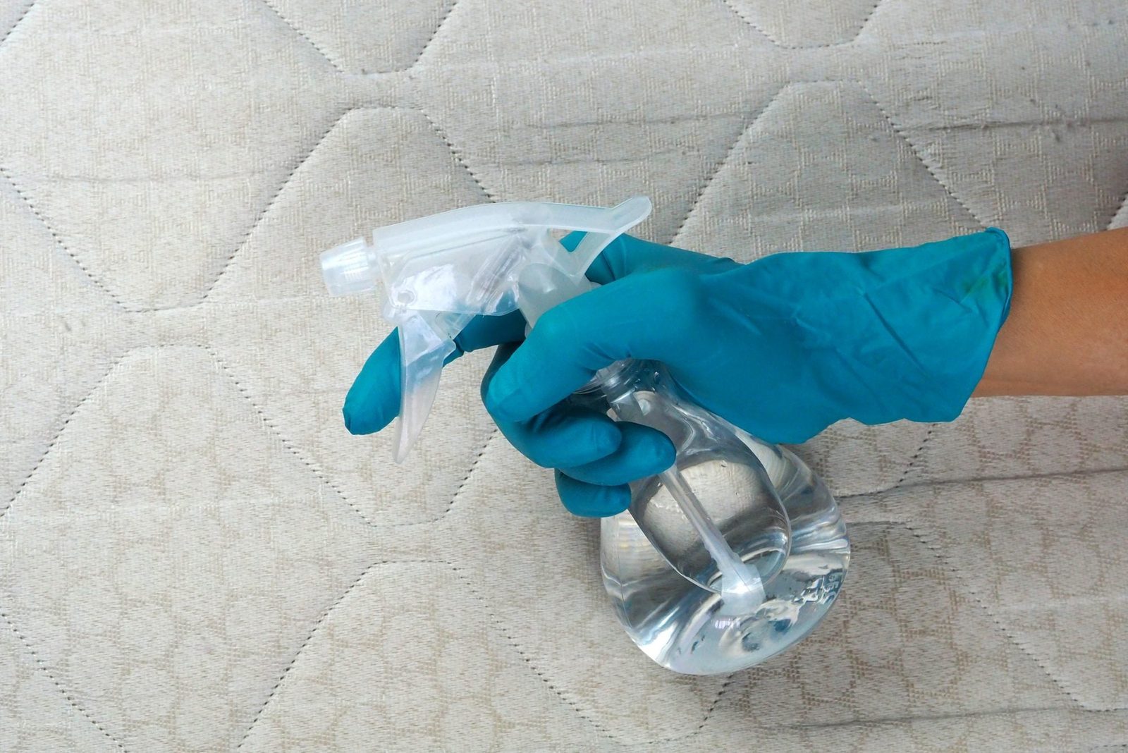 A gloved hand holds a spray bottle, preparing to clean a textured fabric surface, which features a diamond pattern.