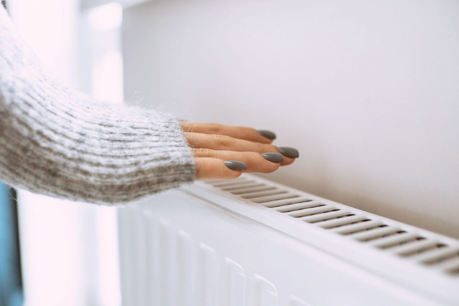 A hand with long, gray nails gently rests on a white radiator, suggesting warmth, against a softly lit indoor background.