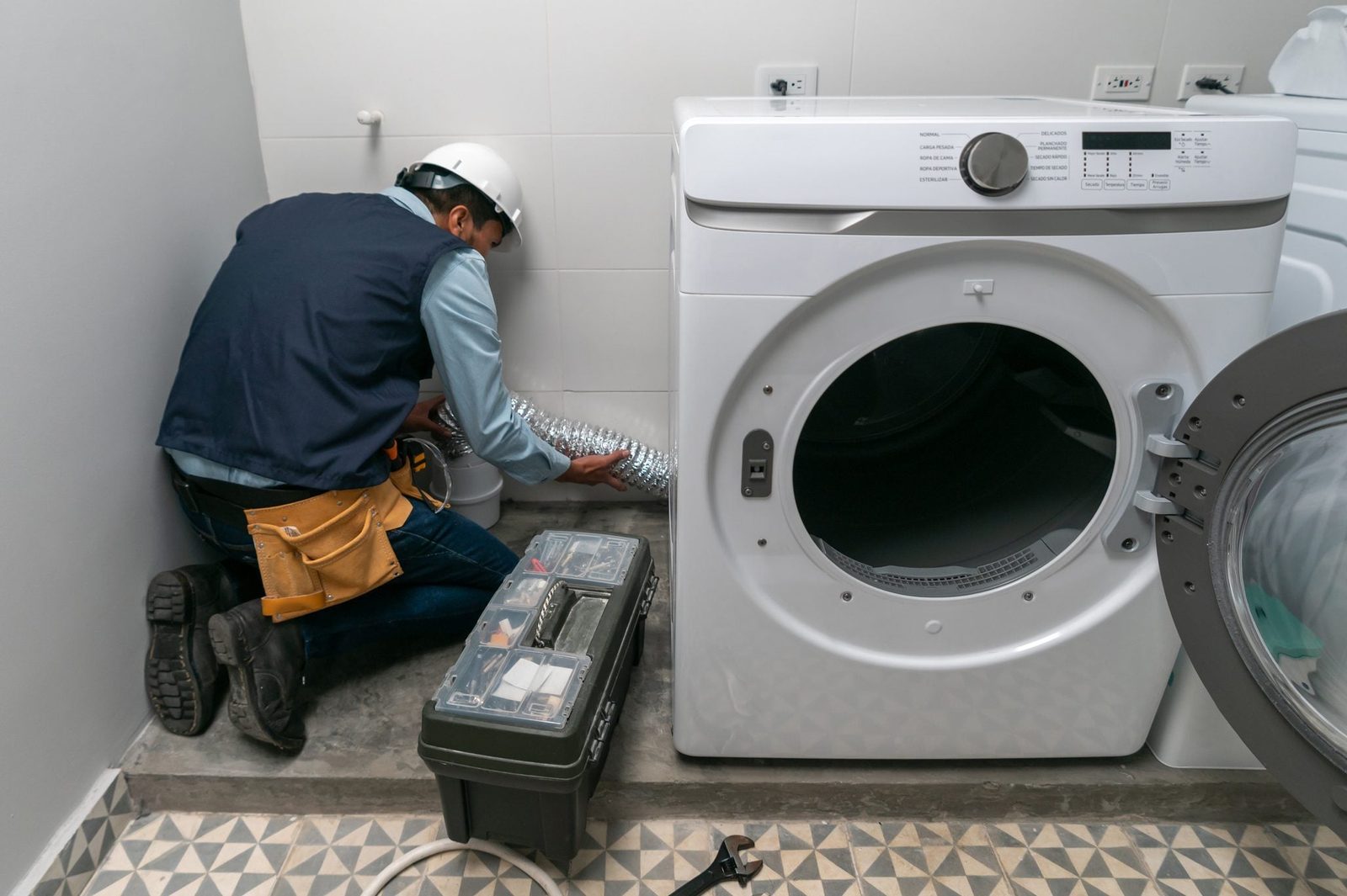 A worker in a helmet connects a duct to a washing machine in a modern laundry room with tiled flooring and a small toolbox nearby.