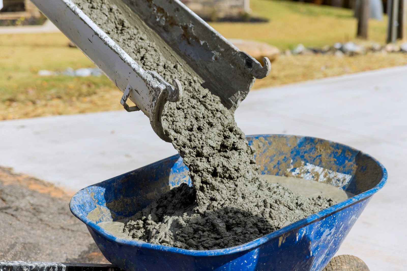 A concrete mixer pours wet concrete into a blue wheelbarrow, positioned on a gravel surface, with a grassy area and paved ground in the back
