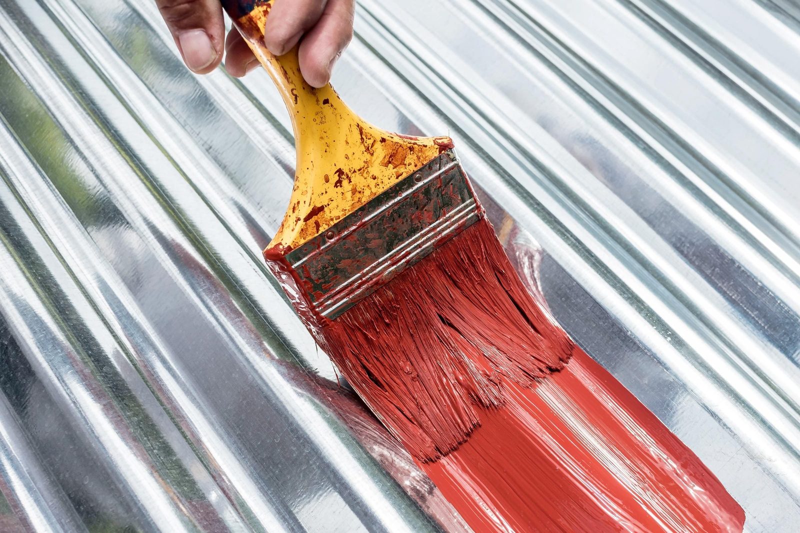 A paintbrush applies red paint onto a shiny, ribbed metal surface, reflecting light from the environment, indicating a painting or restorati