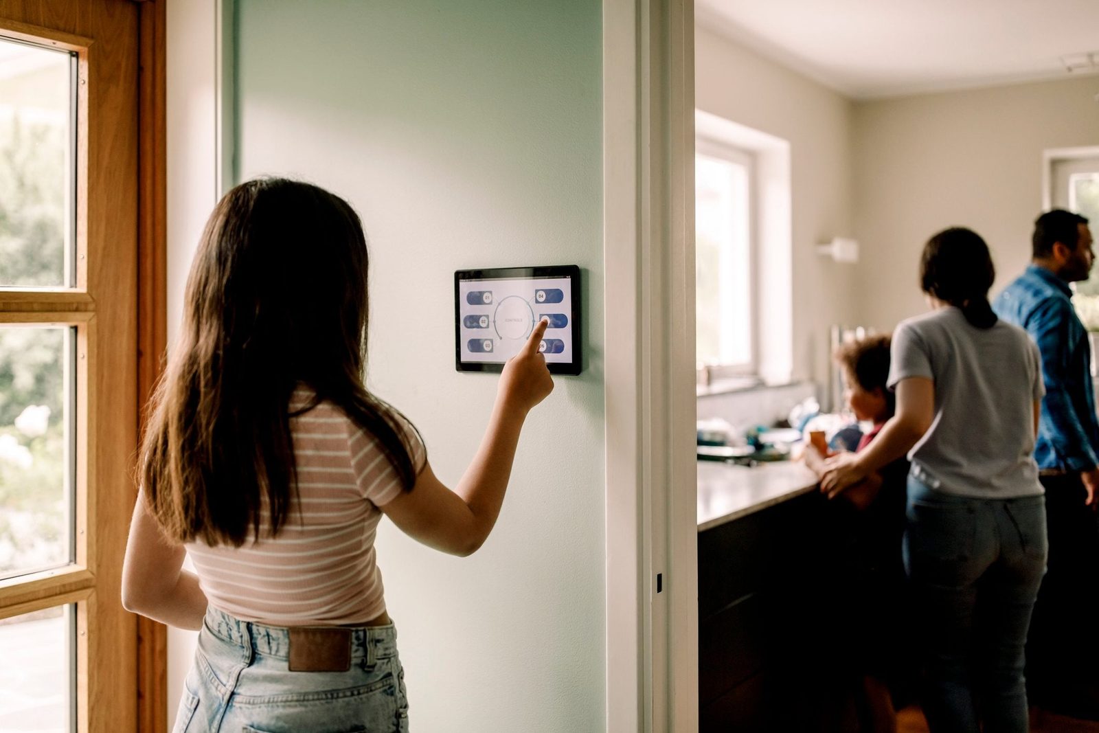 A girl is touching a smart control panel on the wall while two people stand near a kitchen counter in a well-lit room.