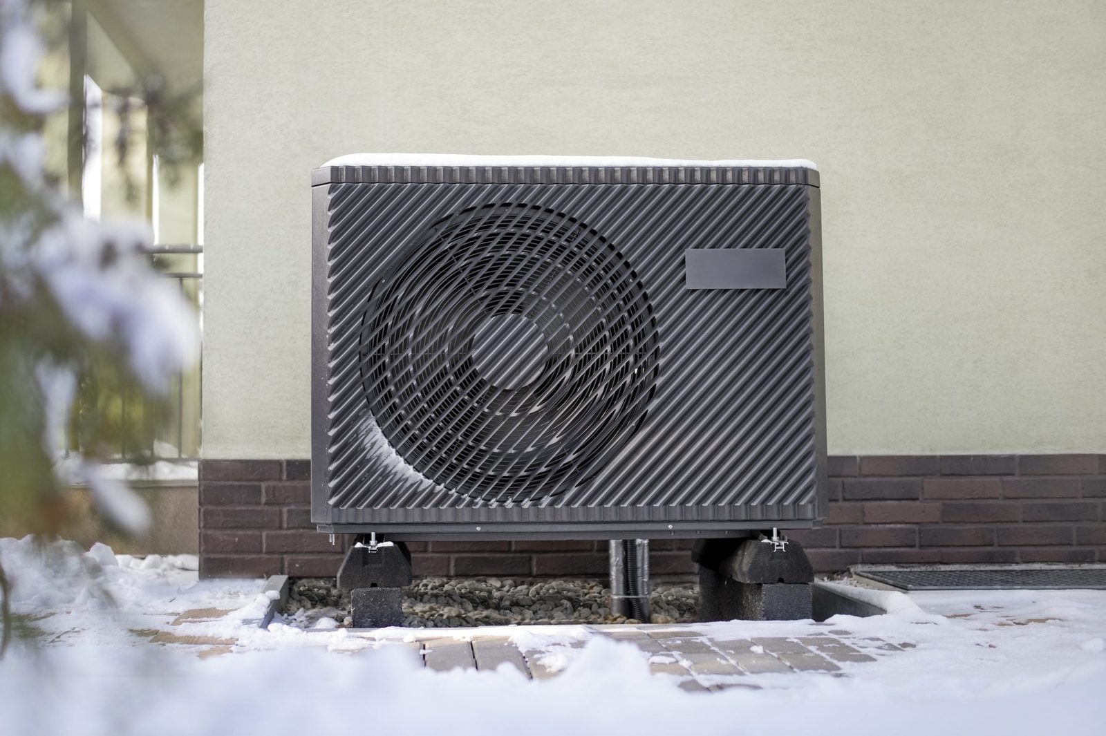 A black outdoor unit is positioned on a snowy pathway beside a house, featuring a circular fan and standing on sturdy blocks against a light