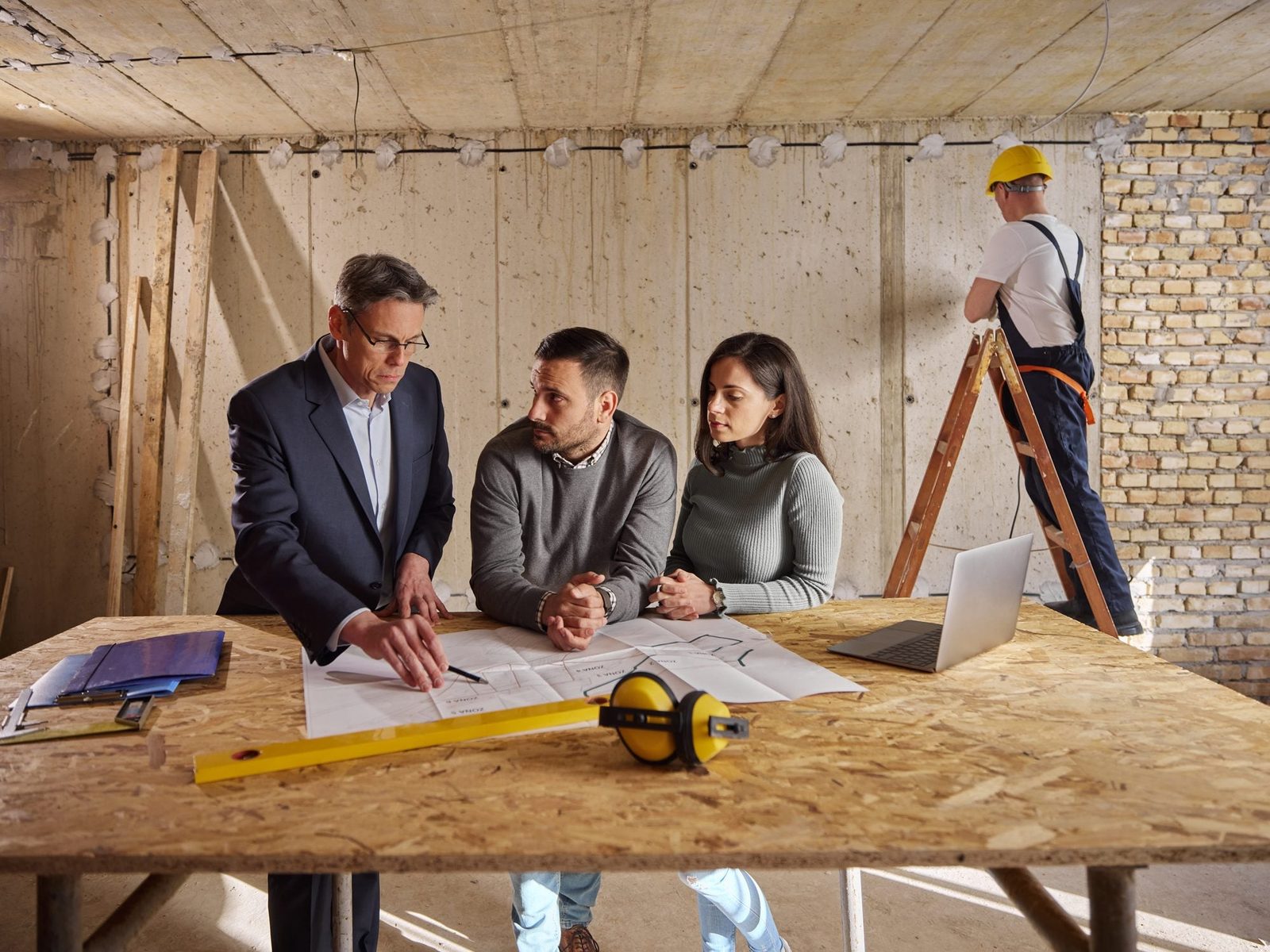 A group discusses building plans at a table, while a worker repairs a wall in a construction site, surrounded by unfinished concrete and woo