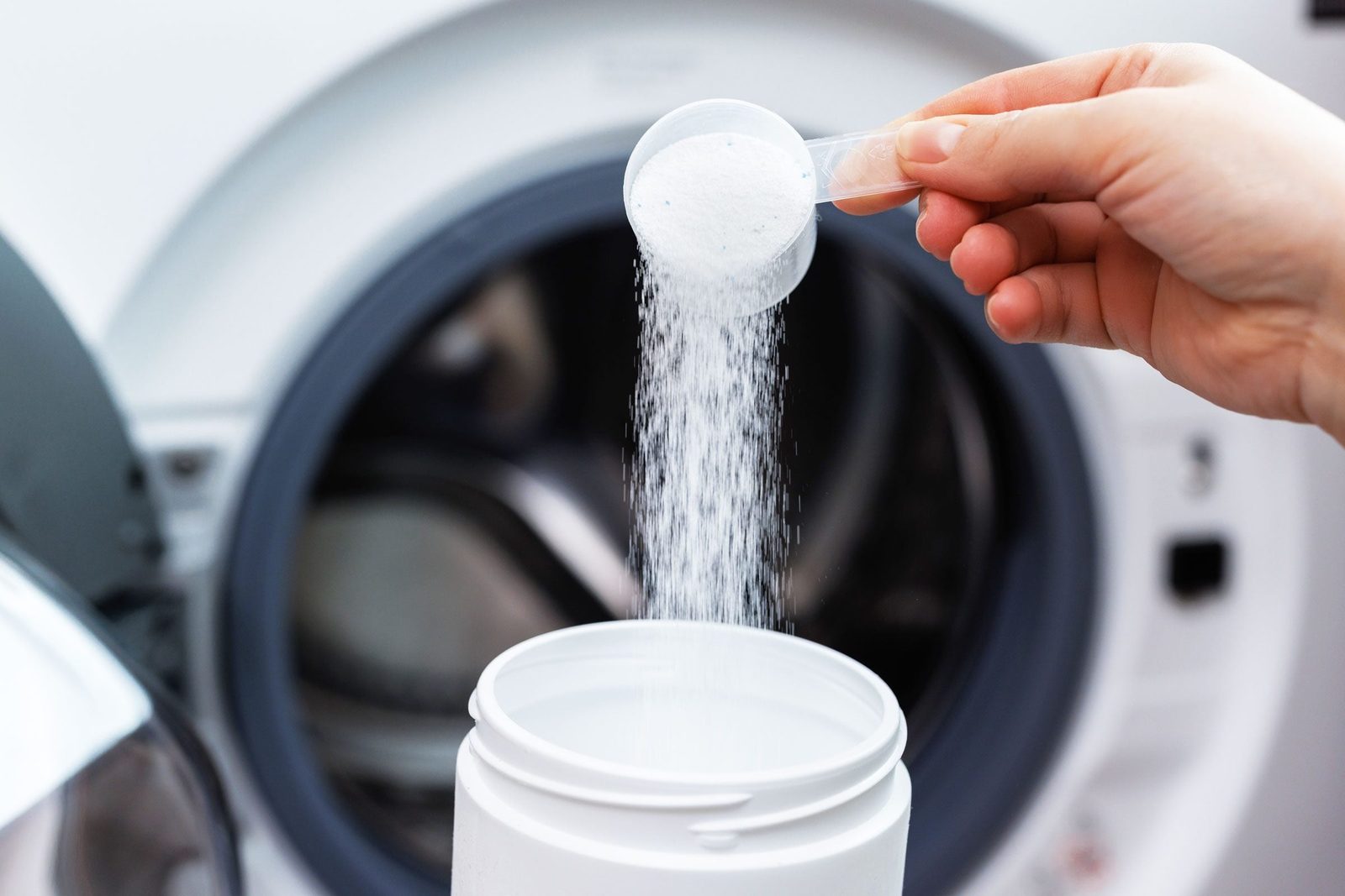 A hand pours laundry detergent from a measuring scoop into a white container, with a washing machine in the background.