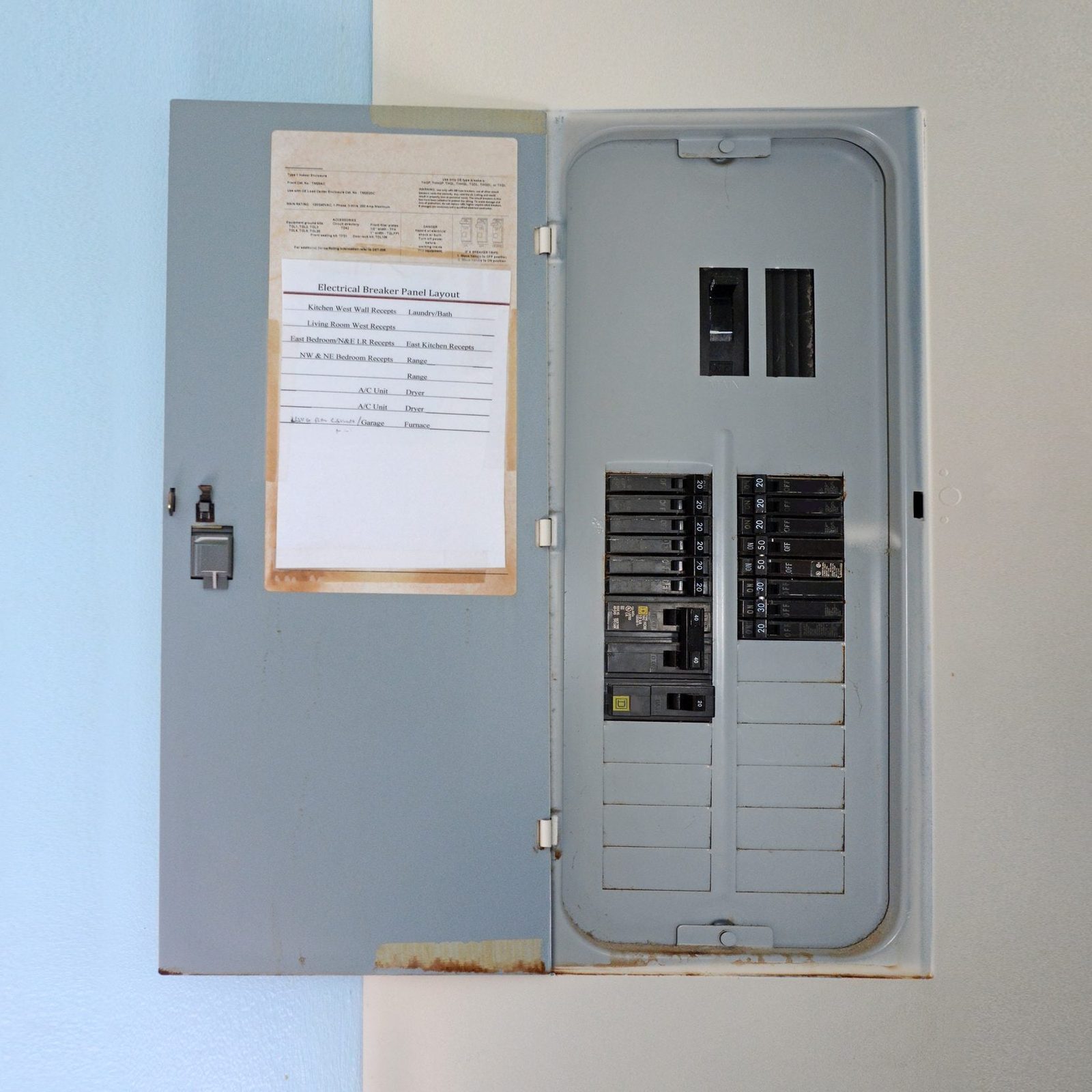 A gray electrical breaker panel with multiple switches is mounted on a wall, featuring a layout guide attached next to it, indicating variou