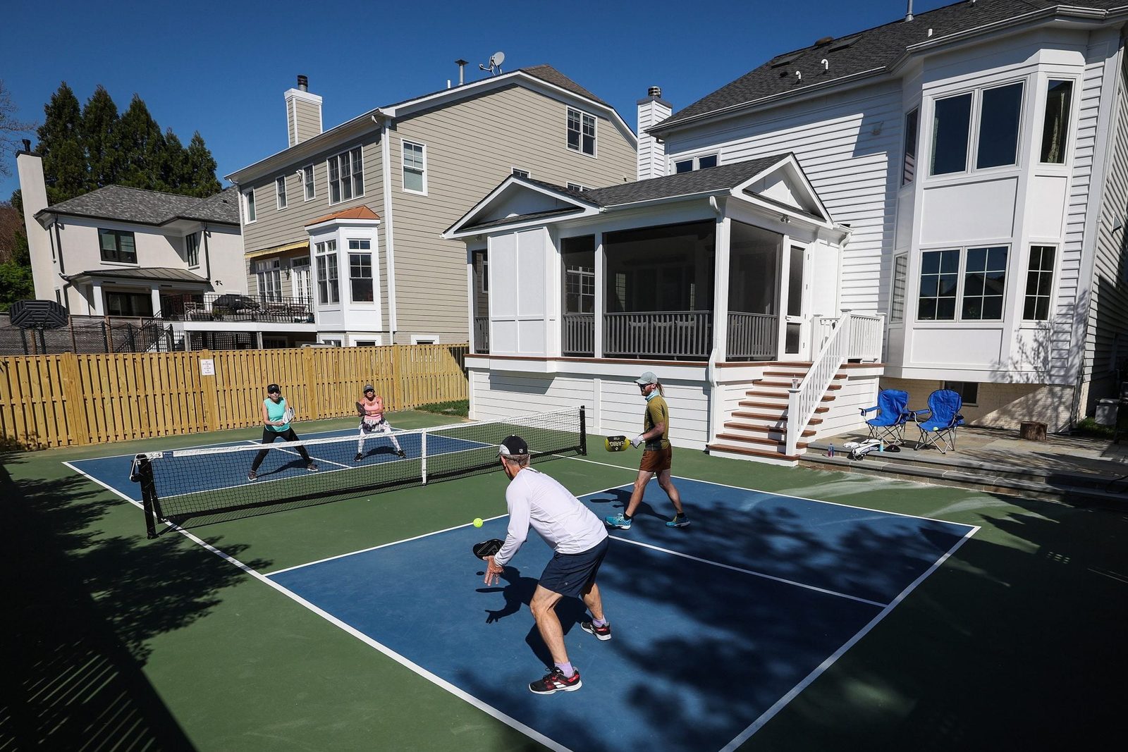 Three people play pickleball on a court in a residential backyard, surrounded by houses and a wooden fence, under a clear blue sky.