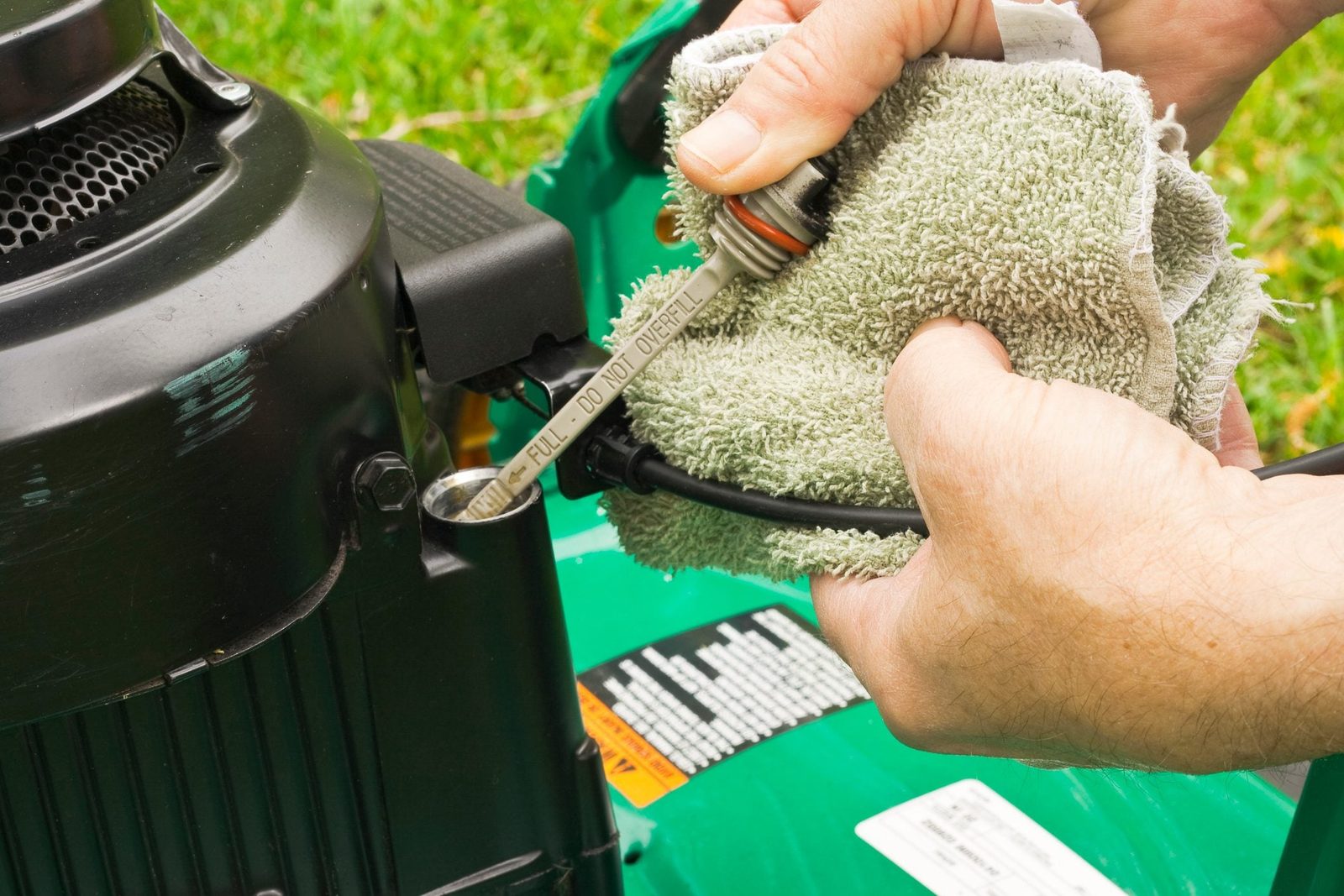 A person checks the oil level in a lawn mower engine using a dipstick while holding a cloth in a grassy outdoor setting.