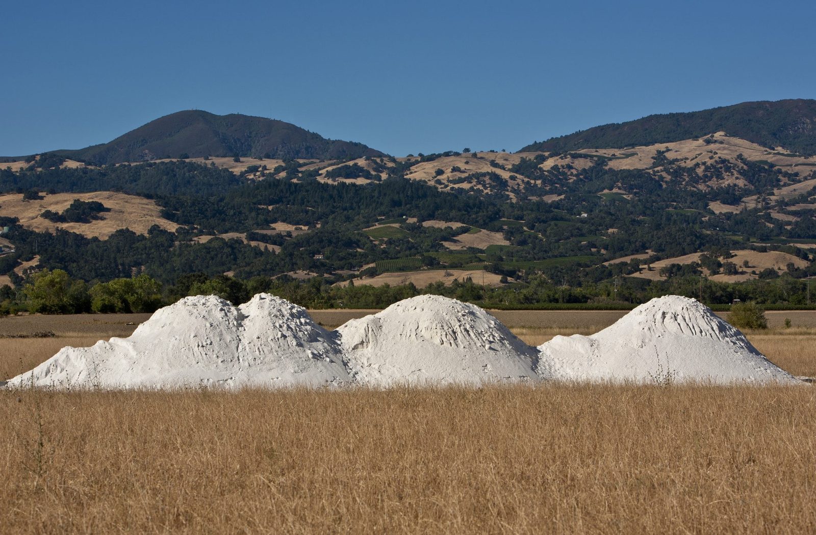 Three large white mounds of material are situated in a dry, golden field, with green hills and a clear blue sky in the background.