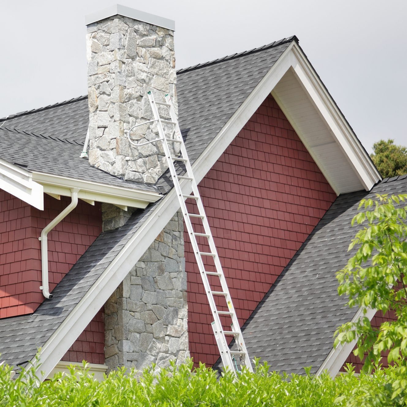 A white ladder leans against a stone chimney on a sloped roof, surrounded by red shingle siding and green foliage below, under a cloudy sky.