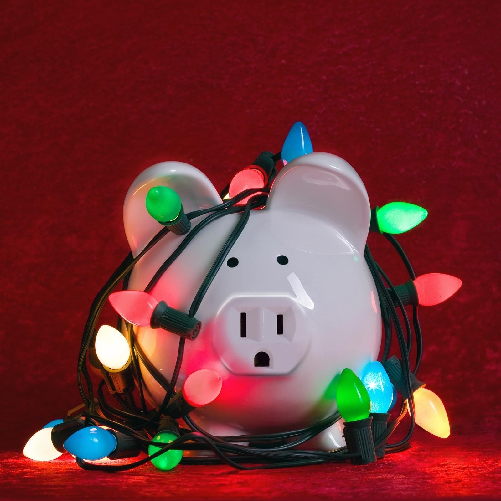 A white piggy bank is entangled in colorful Christmas lights against a red backdrop, suggesting a festive but chaotic atmosphere.