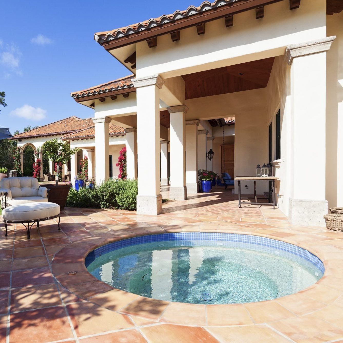 A round hot tub sits on a tiled patio adorned with vibrant flowers, beside a spacious, sunlit house featuring arched columns and a terracott