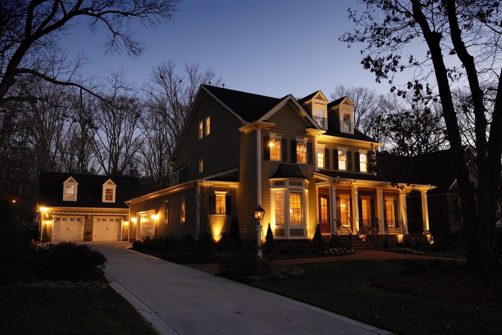 A well-lit two-story house stands along a driveway, surrounded by trees, as dusk settles in. Soft lighting highlights its features and lands