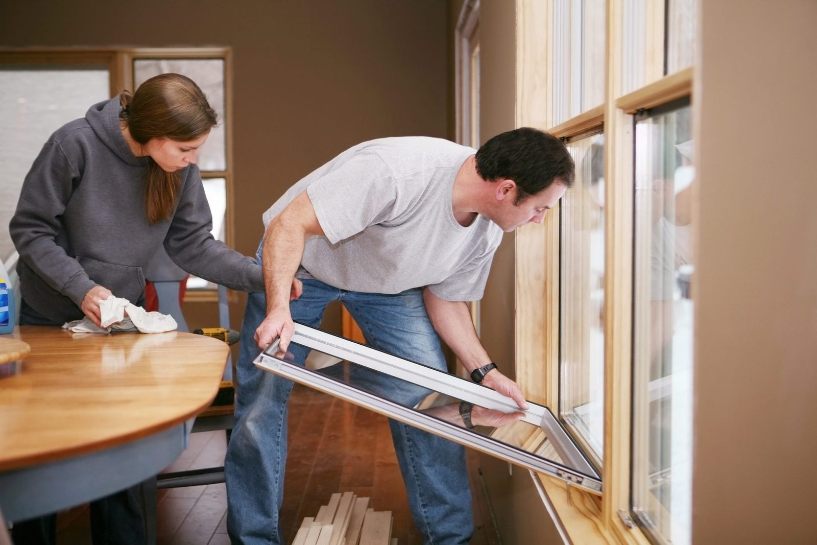Two people work together to install a window frame in a room with wooden floors and light-colored walls, focusing intently on their task.
