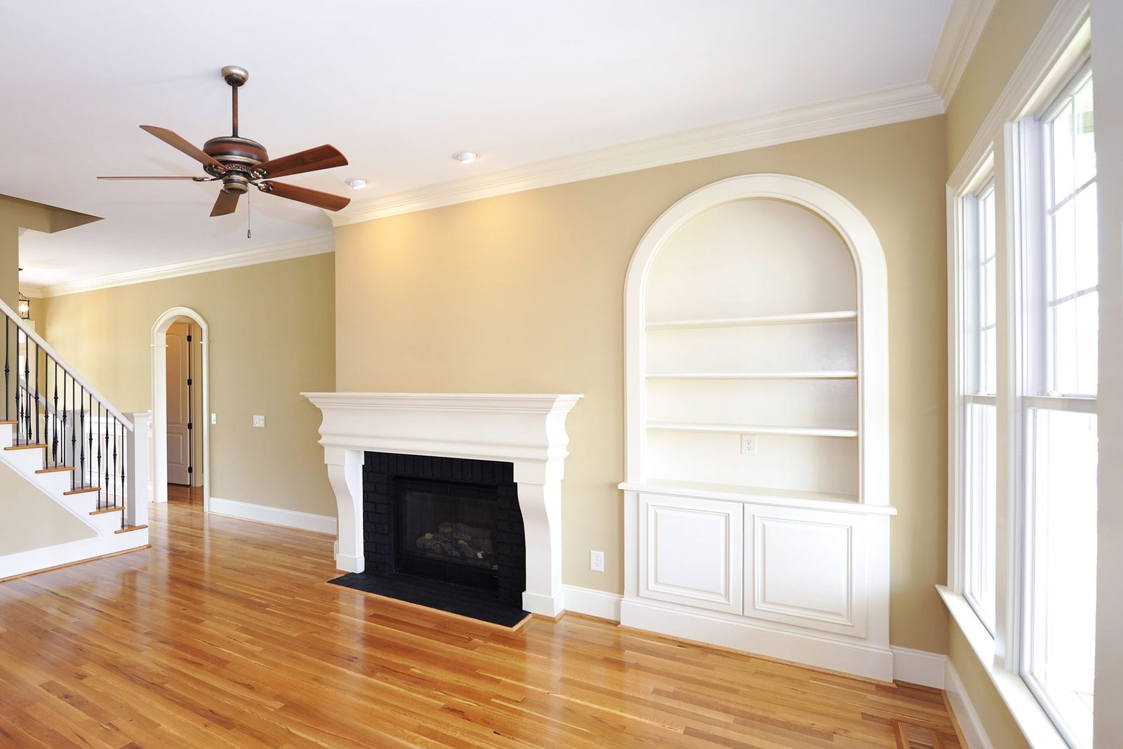 A ceiling fan spins above a wooden floor, while a white fireplace and built-in shelves enhance the light-colored walls of a spacious living