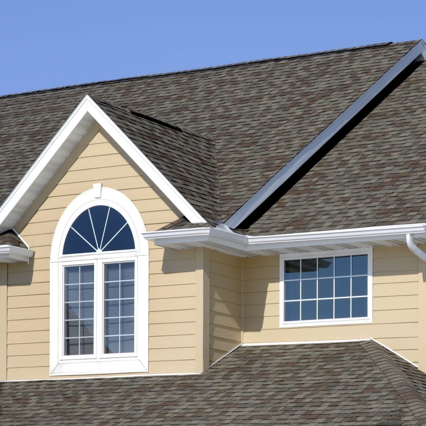 A house's upper exterior featuring a sloped roof, two windows, and beige siding under a clear blue sky.
