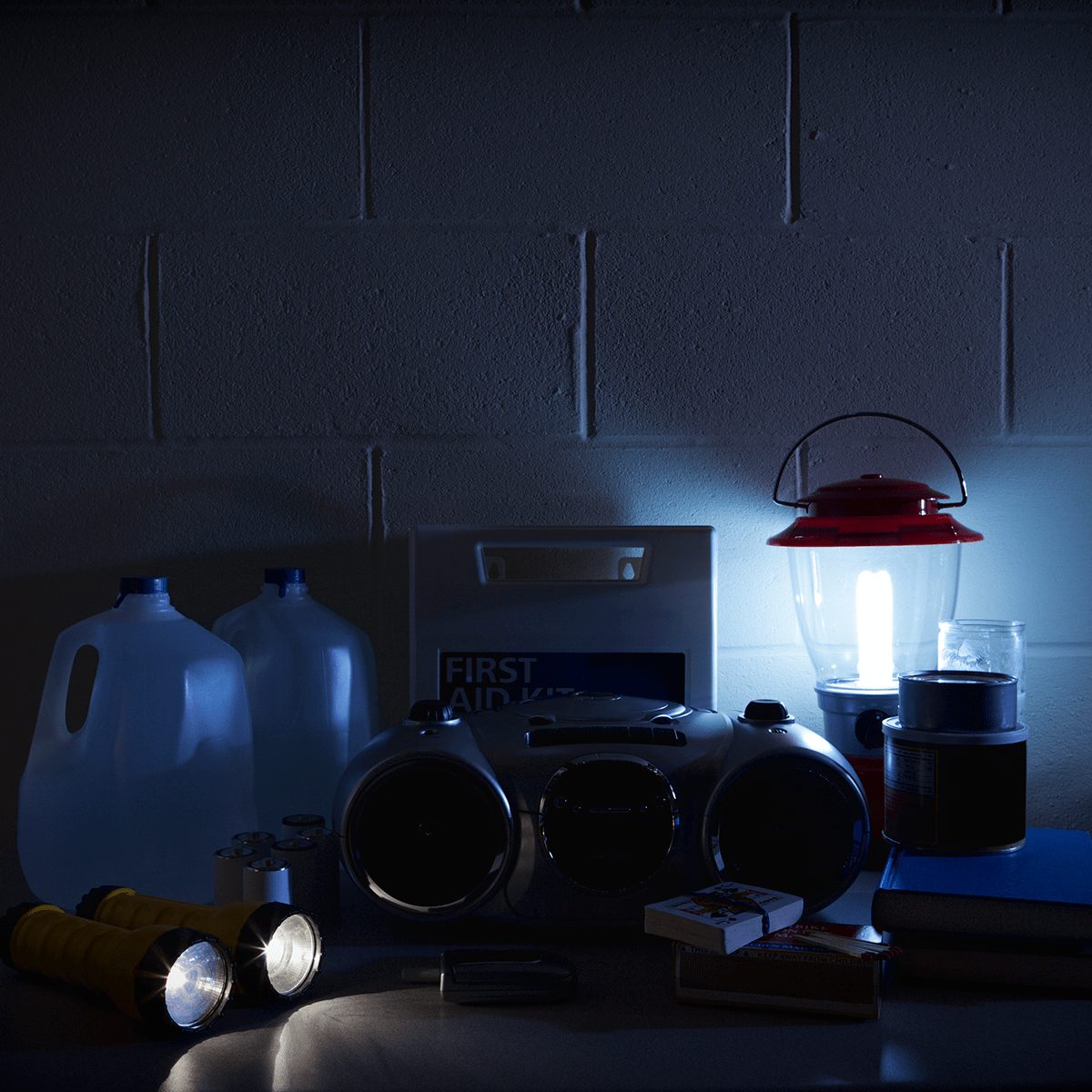 A first aid kit, lantern, bottles of water, and flashlights are arranged on a table in a dimly lit, indoor environment.