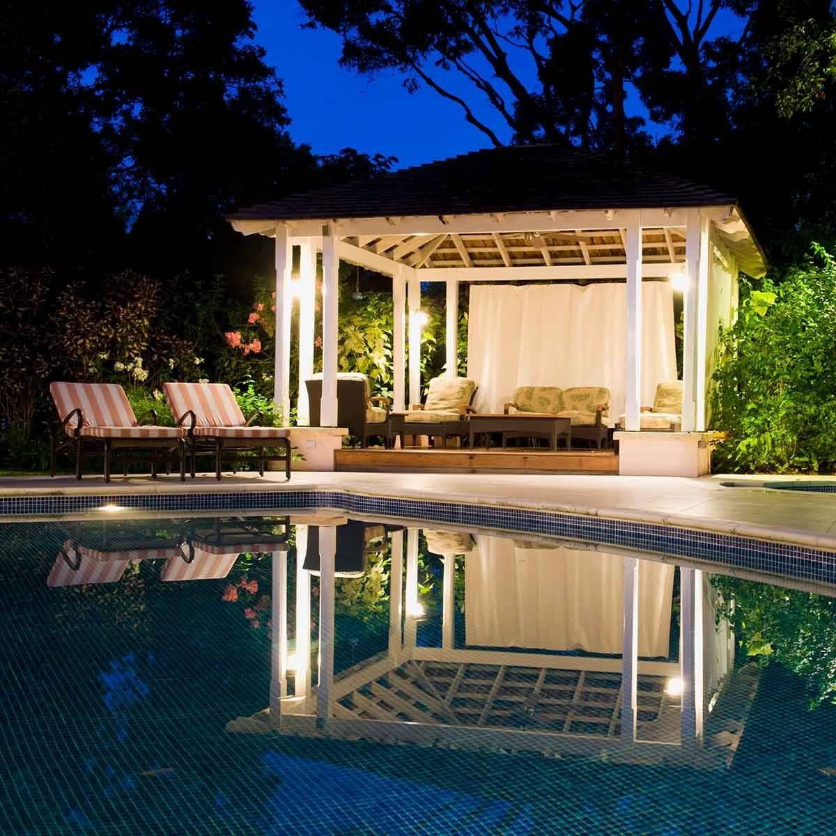 A gazebo stands illuminated at night beside a swimming pool, reflecting in the water, surrounded by greenery and two striped lounge chairs.