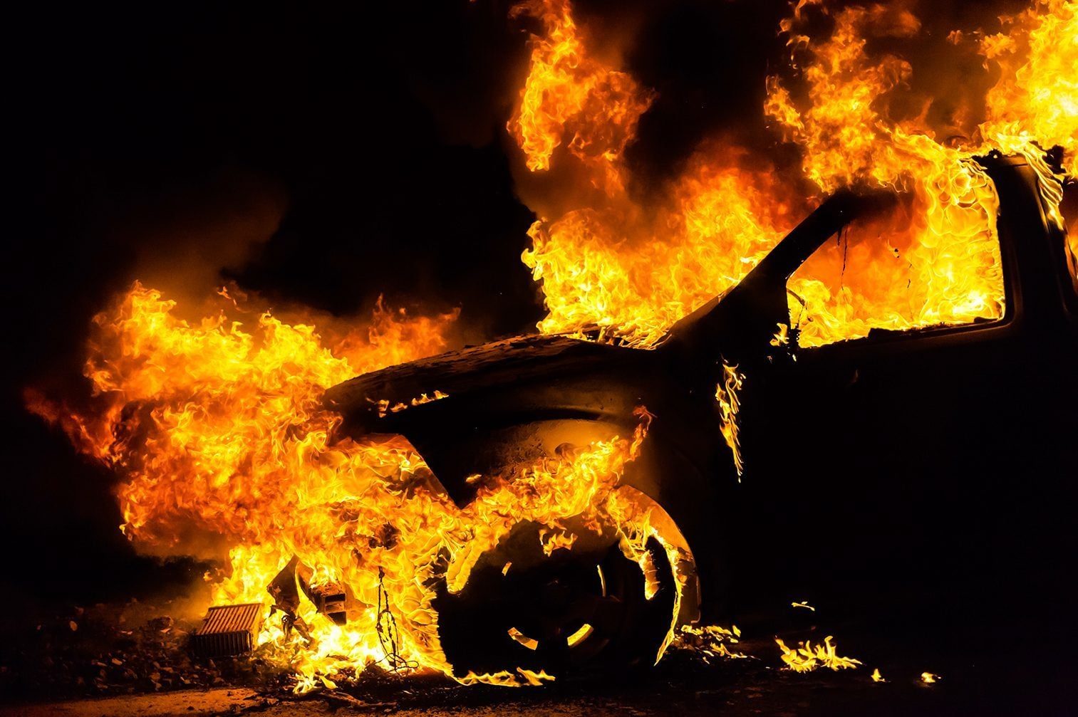 A car is engulfed in flames, burning intensely in a dark environment, with bright orange and yellow flames consuming its structure.
