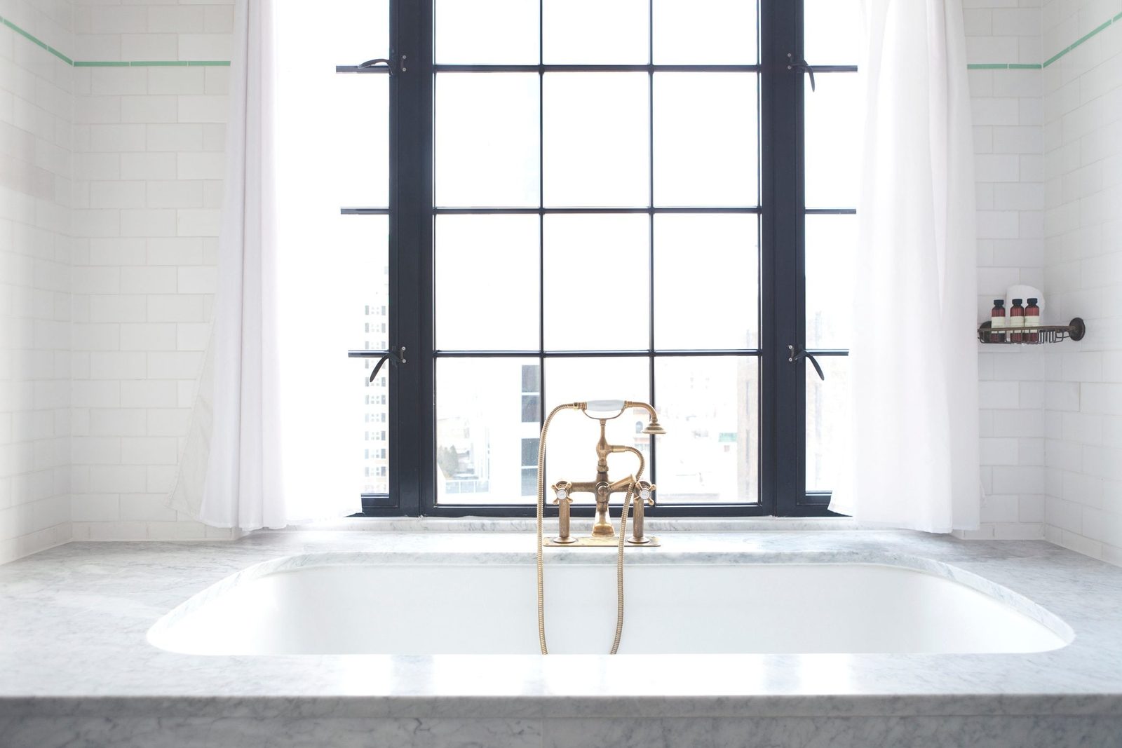 A gold faucet pours water into a white bathtub, surrounded by marble edges and large windows covered with sheer curtains, allowing natural l