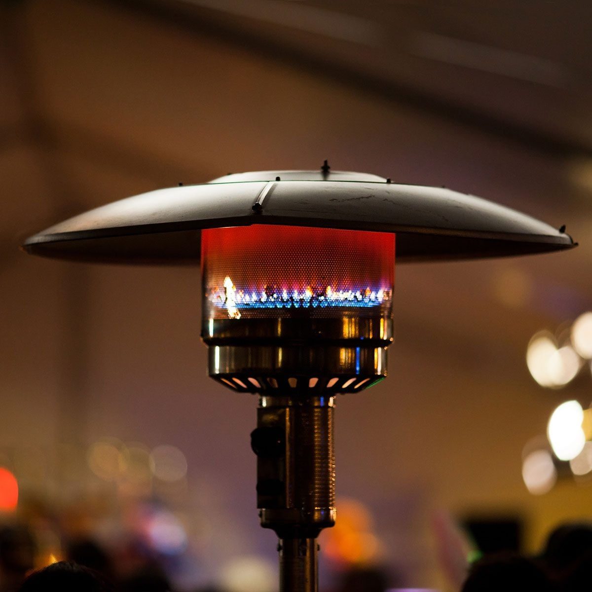 A gas patio heater emits bright flames and heat, standing amidst a blurred indoor crowd with soft lighting in the background.