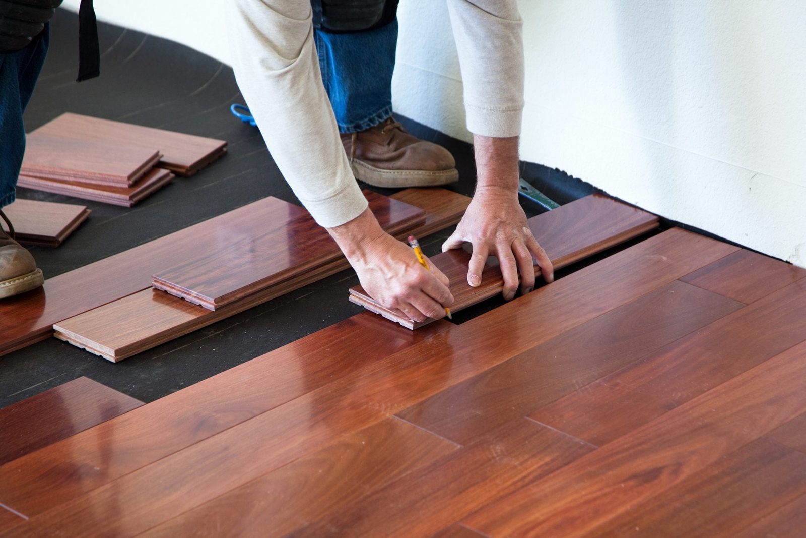 A person is marking wooden flooring planks on a black underlayment within a room, preparing to install them against a light-colored wall.