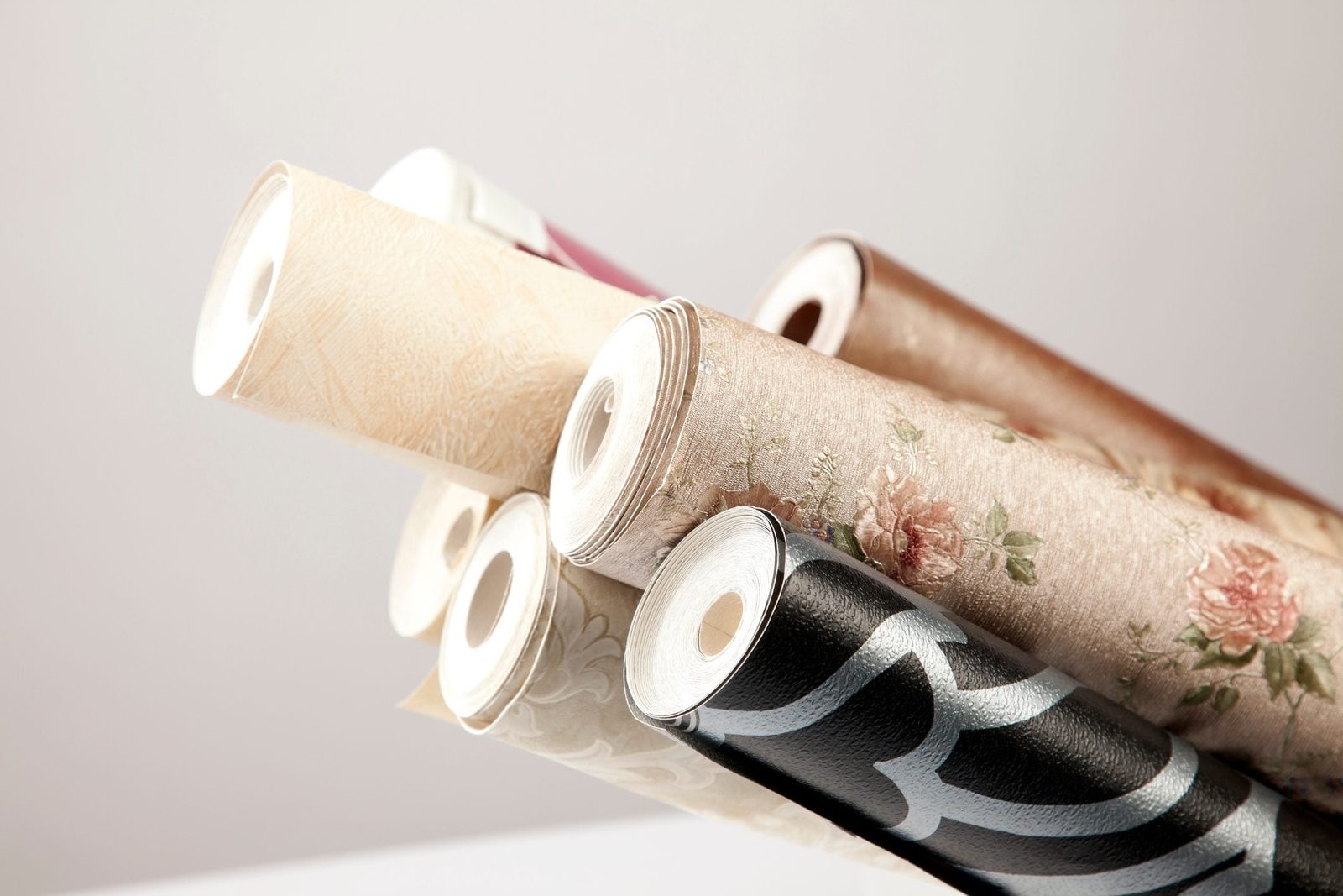 Rolls of decorative wallpaper lean against each other, showcasing various patterns and colors, in a minimalistic, well-lit environment.