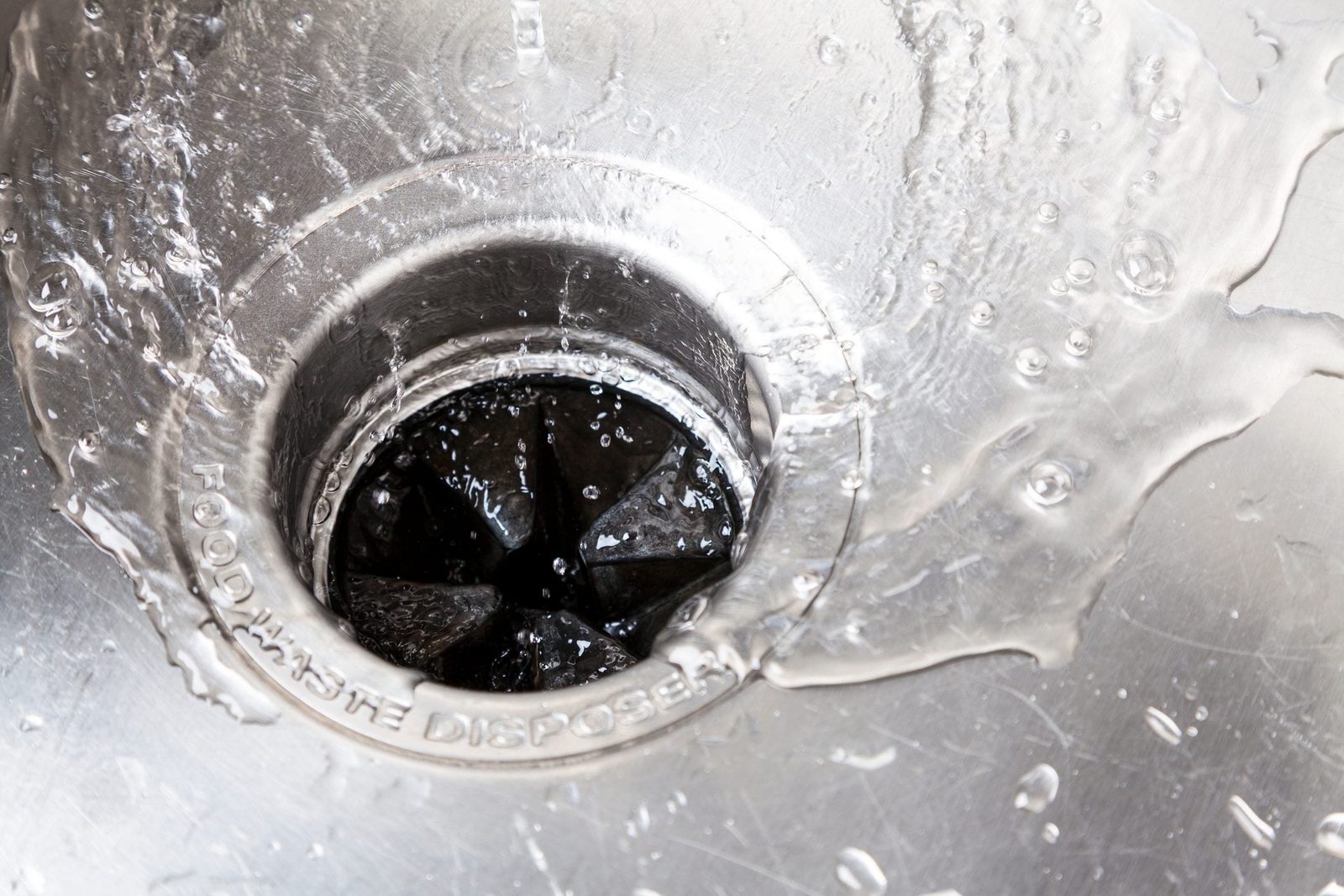 Water flows into a stainless steel sink's drain, creating splashes around the edges, while the interior features a food waste disposal unit.
