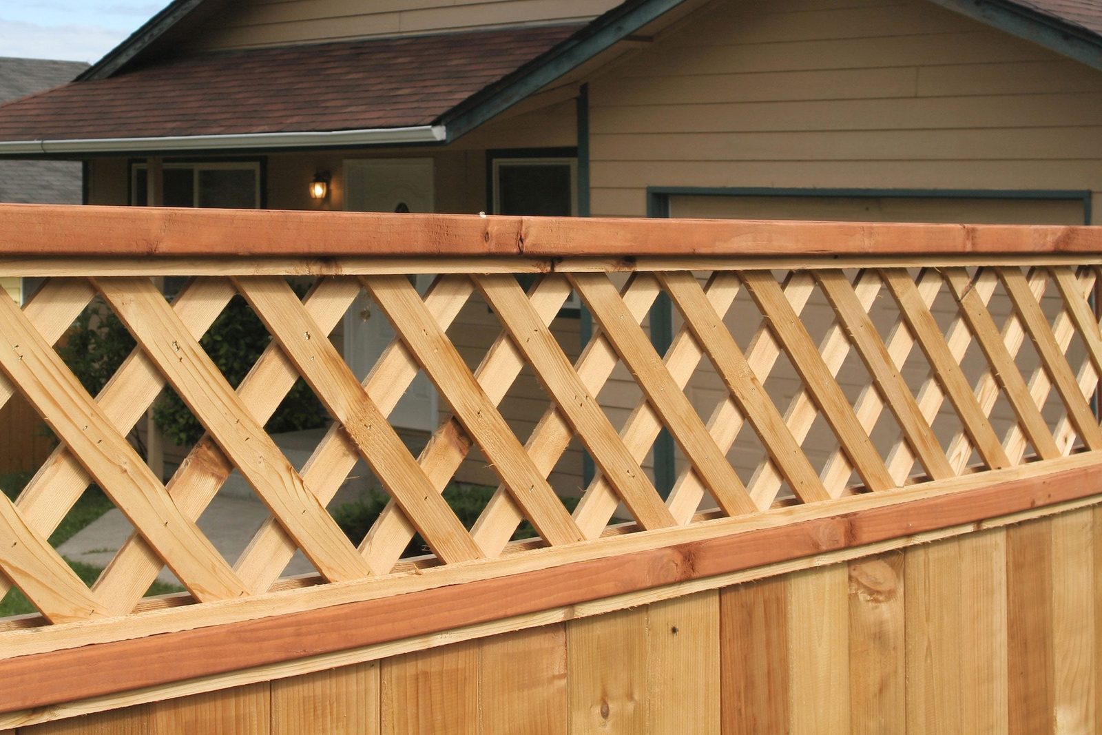 A wooden fence with a lattice top defines a boundary, positioned in front of a house with a roof and porch, surrounded by grass.