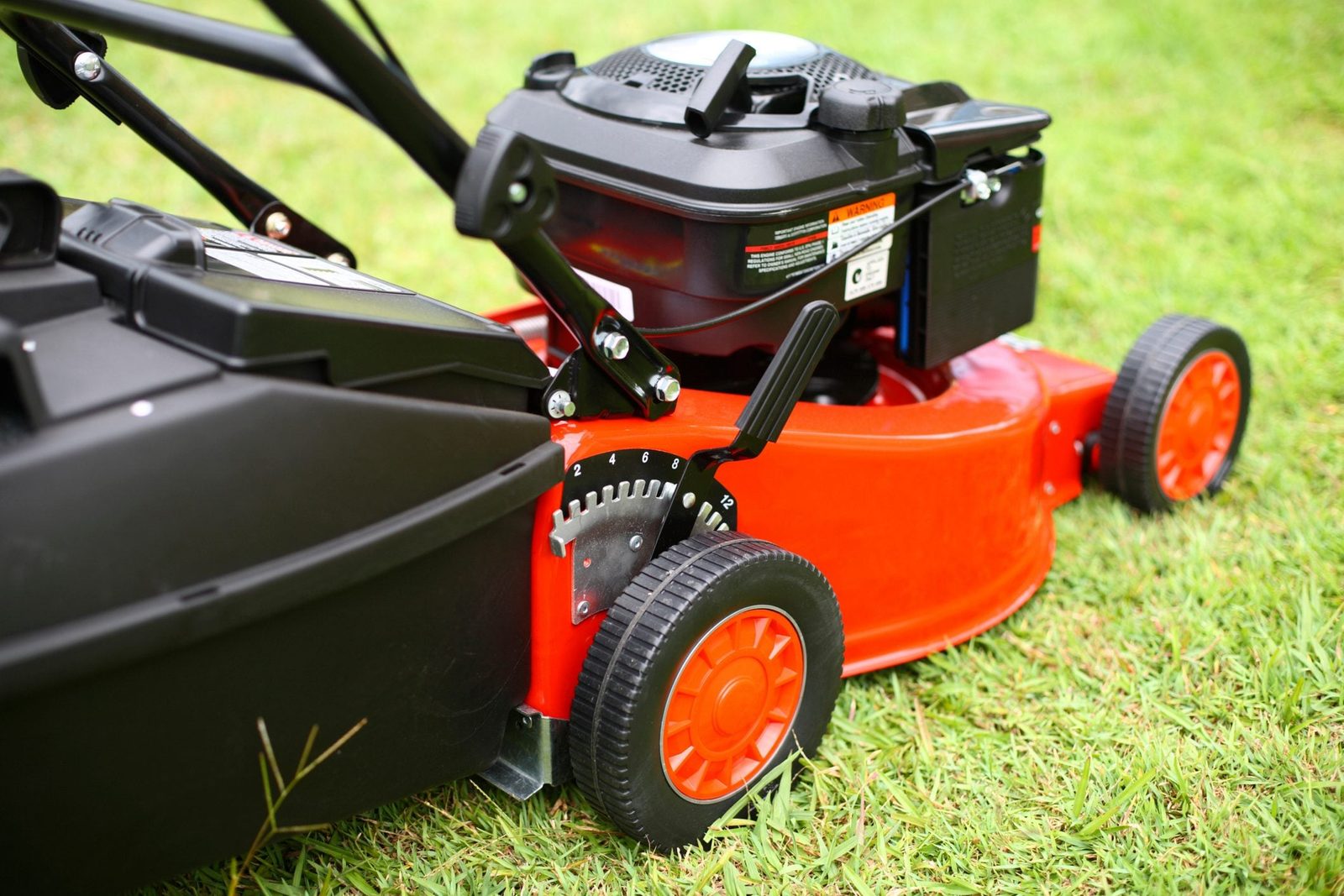 A lawn mower rests on green grass, with its engine visible and a lever for height adjustment beside its right wheel.