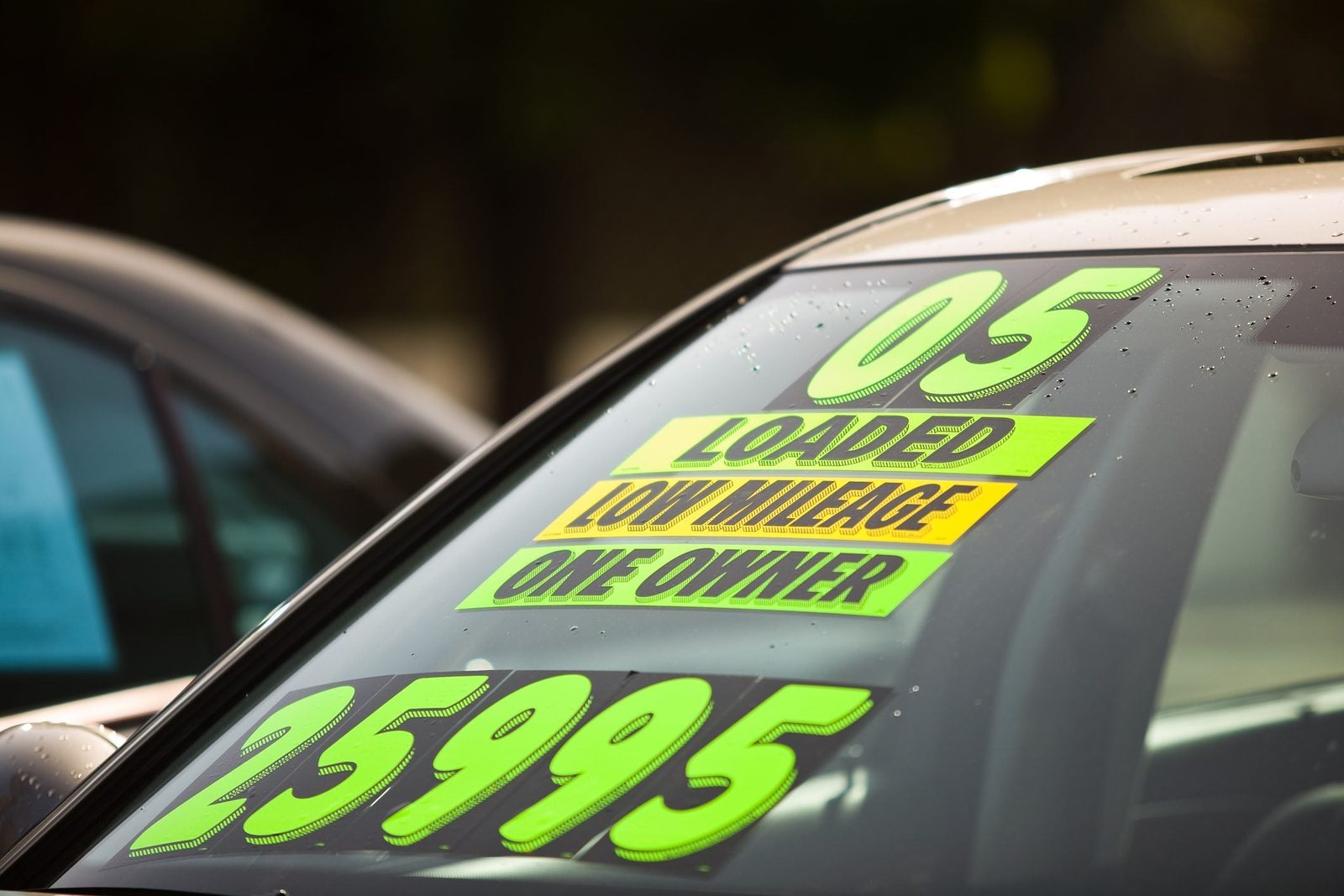 A car features a brightly colored sales sticker displaying "05," "LOADED," "LOW MILEAGE," "ONE OWNER," and a price of "25995" against a blur
