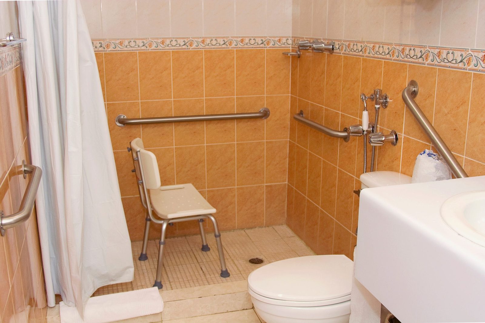 A shower chair sits near a toilet in a small bathroom, featuring tiled orange walls and grab bars for support, with a shower curtain partial