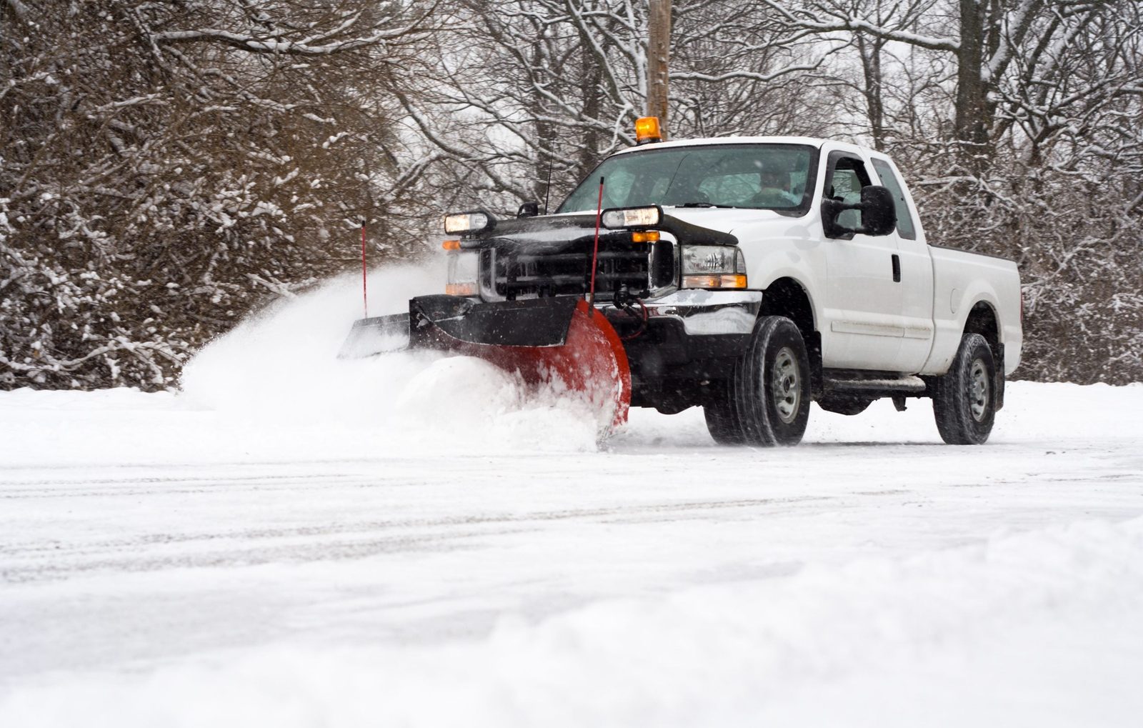 A white pickup truck with a snow plow clears snow from a snowy road surrounded by trees in a winter landscape.