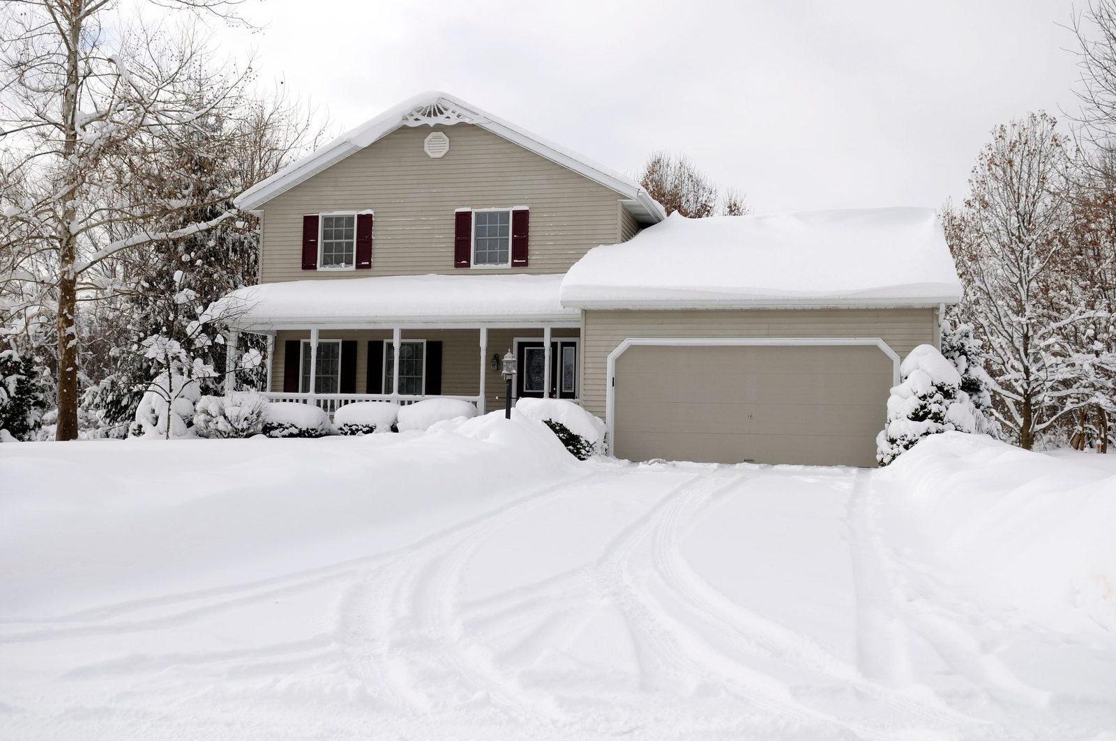 A two-story house is partially covered in snow, with a snow-covered driveway leading to an attached garage, surrounded by snowy trees and sh
