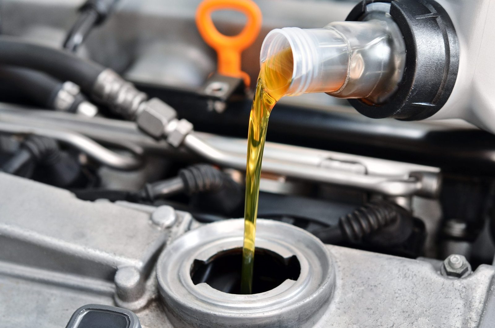 A clear container pours golden oil into an engine’s oil fill opening, surrounded by metal machinery and rubber hoses in a mechanical environ