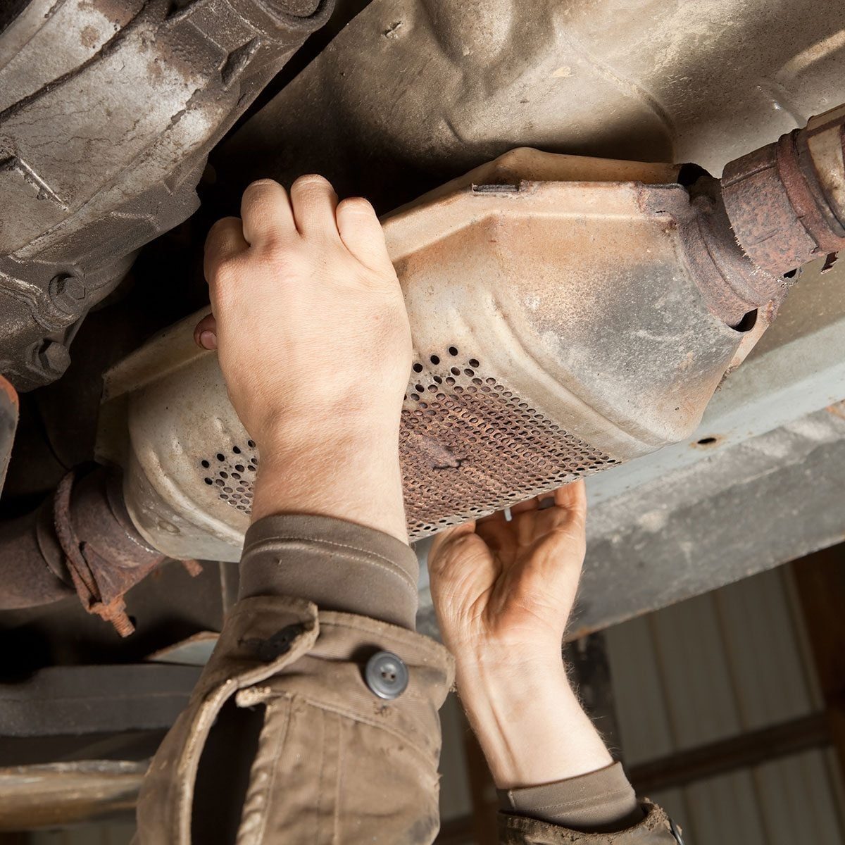 A person is installing a catalytic converter beneath a vehicle, using both hands to position it in a maintenance garage setting.