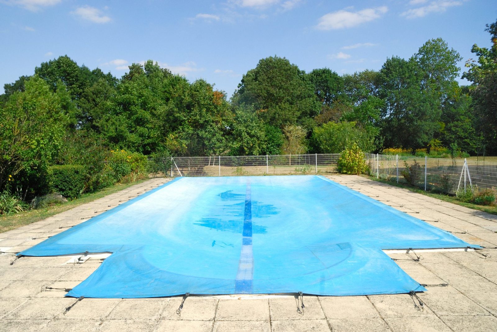 A blue pool cover rests over an empty swimming pool, surrounded by a concrete deck and lush greenery under a clear blue sky.