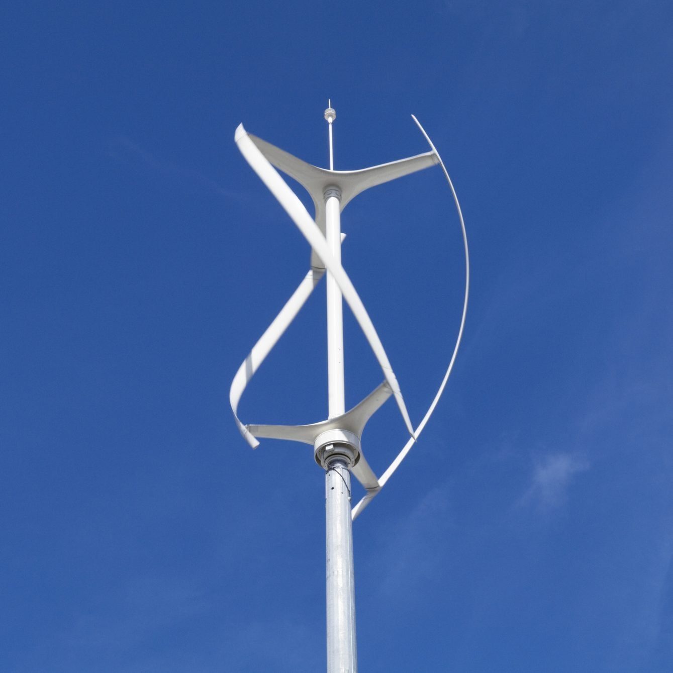 A vertical wind turbine spins, harnessing wind energy against a clear blue sky, mounted on a tall metallic pole.