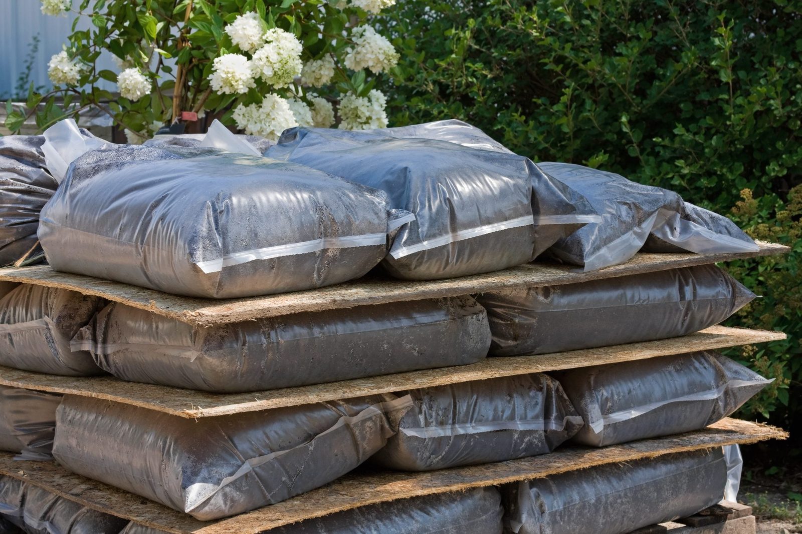 Plastic bags filled with dark material are stacked on wooden pallets, surrounded by greenery and white flowering plants in a bright outdoor