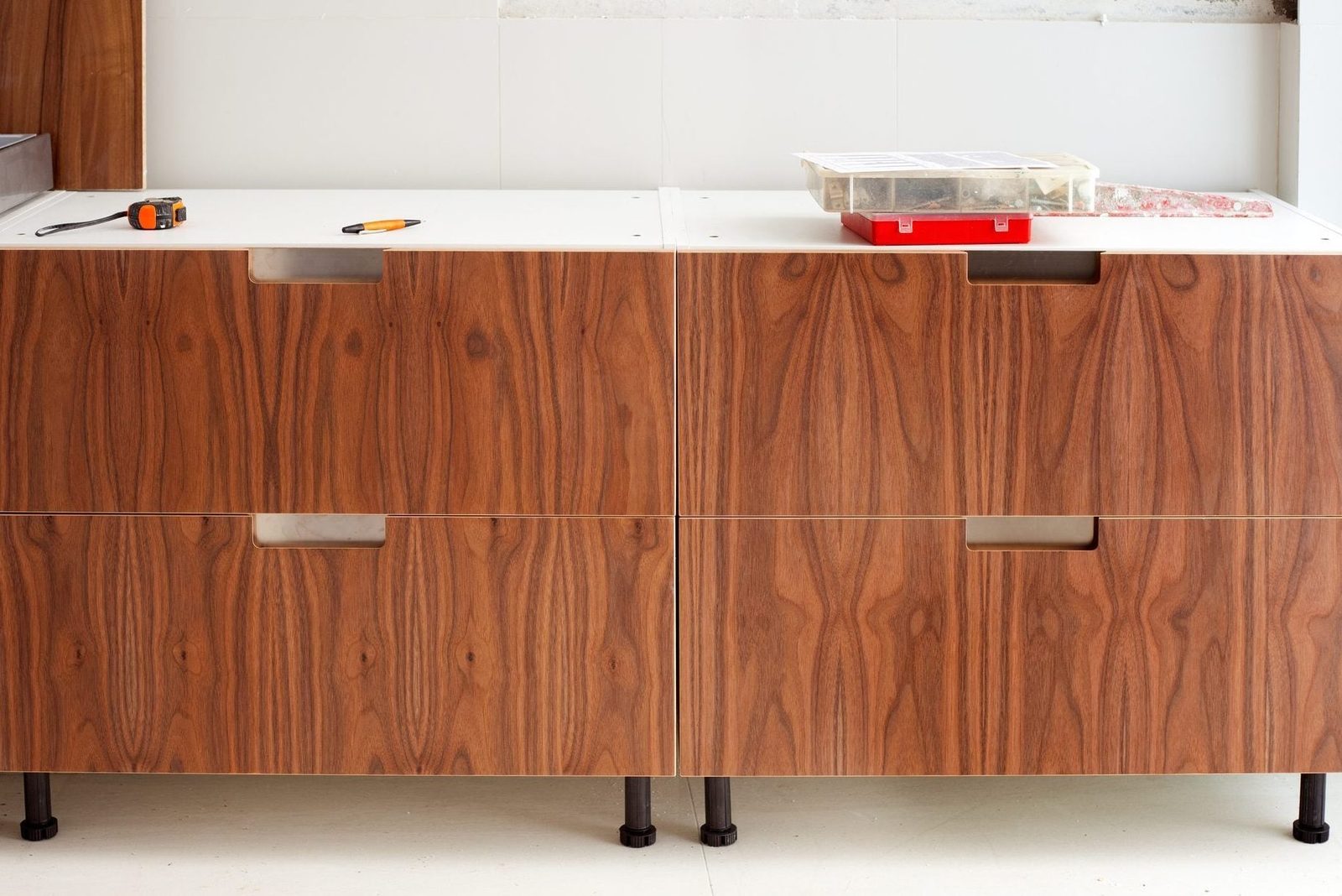 A wooden cabinet with two drawers is displayed, equipped with a measuring tape and tools, in a bright, unfinished room with light-colored wa