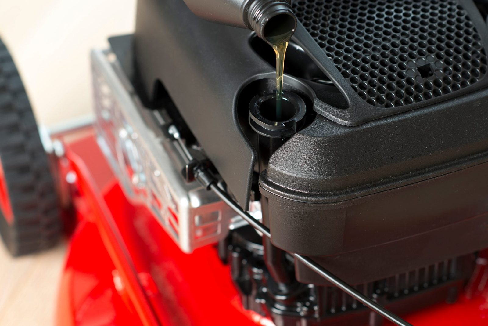 A lawnmower engine is being filled with oil from a black bottle, situated on a red base, with a textured cover nearby.