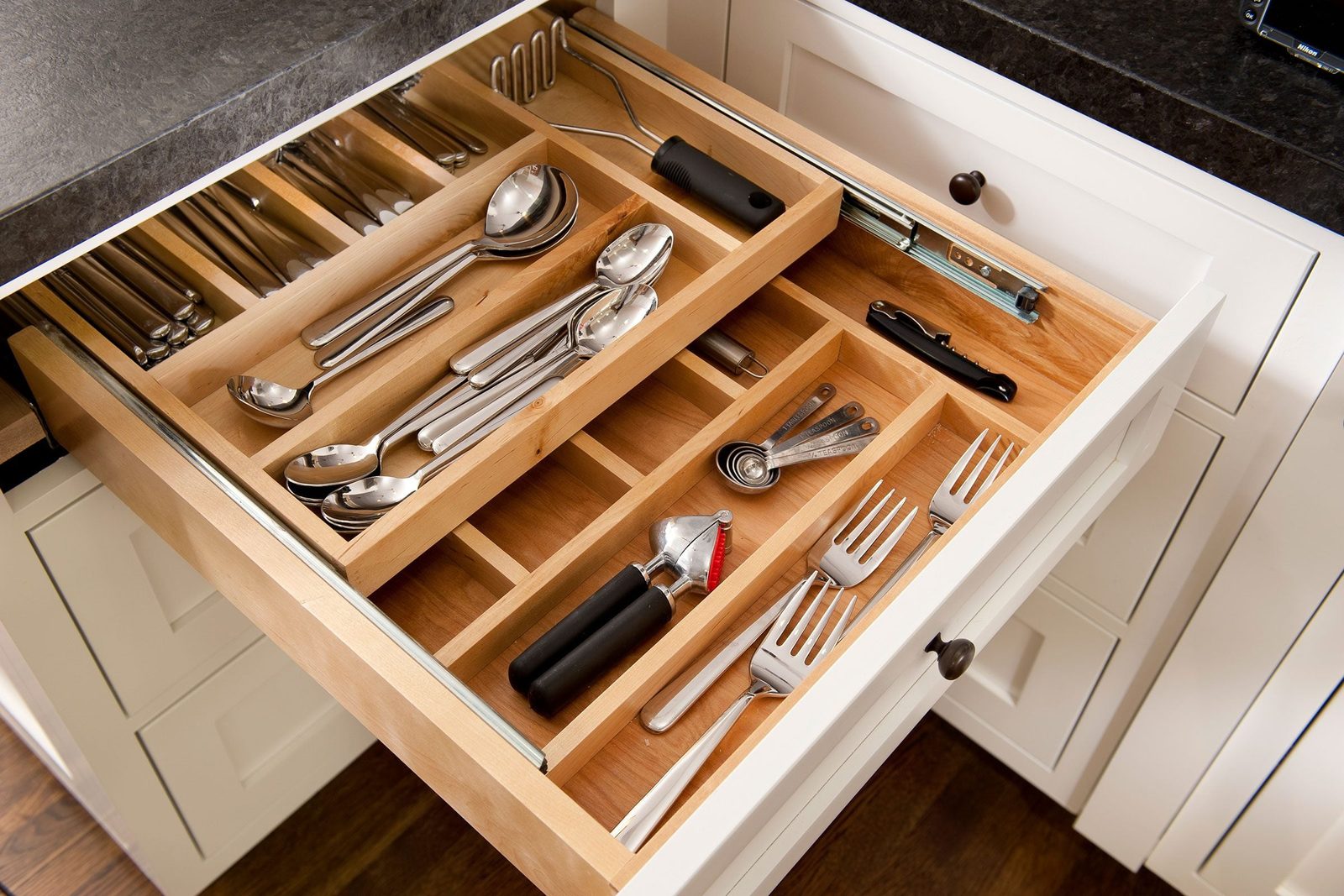 A kitchen drawer filled with organized utensils, including spoons, forks, and measuring tools, rests under a dark countertop in a modern kit