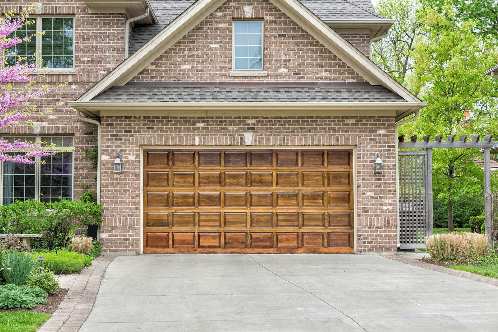A wooden garage door stands closed on a paved driveway, bordered by green plants, a brick house, and a purple tree. Lush greenery surrounds