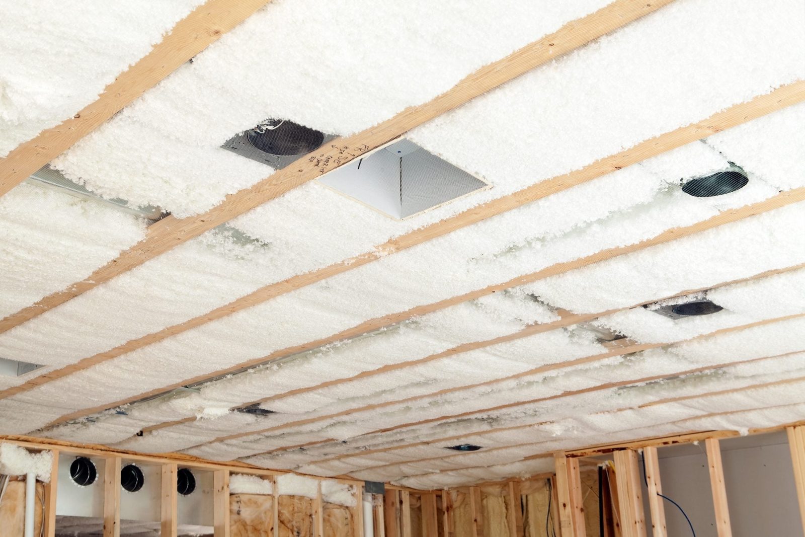 Insulation material covers a wooden ceiling with exposed beams, featuring square openings for lighting fixtures, in an unfinished room under