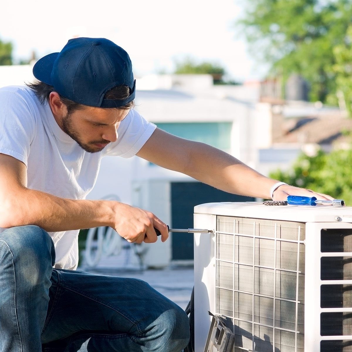 A man kneels, using a screwdriver to repair an air conditioning unit on a rooftop, surrounded by buildings and greenery under a clear sky.