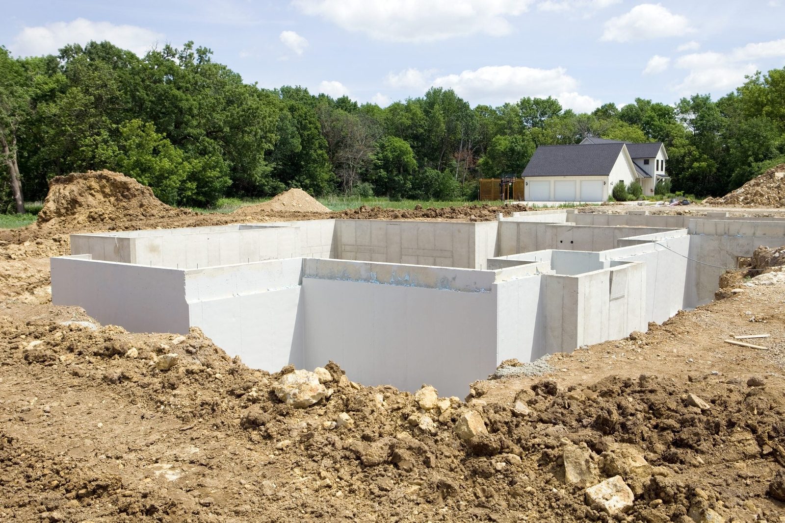 A concrete foundation is situated in an excavated area, surrounded by dirt piles and green trees, with a house visible in the background.