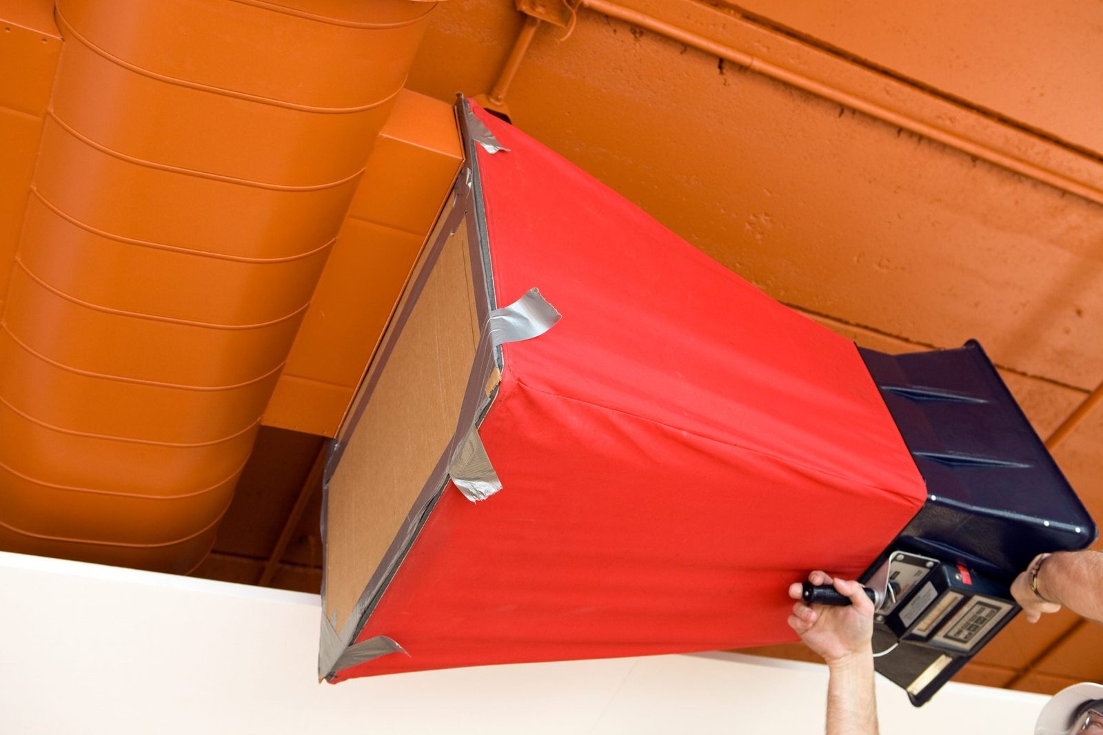 A person is adjusting a red and black object attached to the ceiling, surrounded by orange pipes and industrial-style lighting.