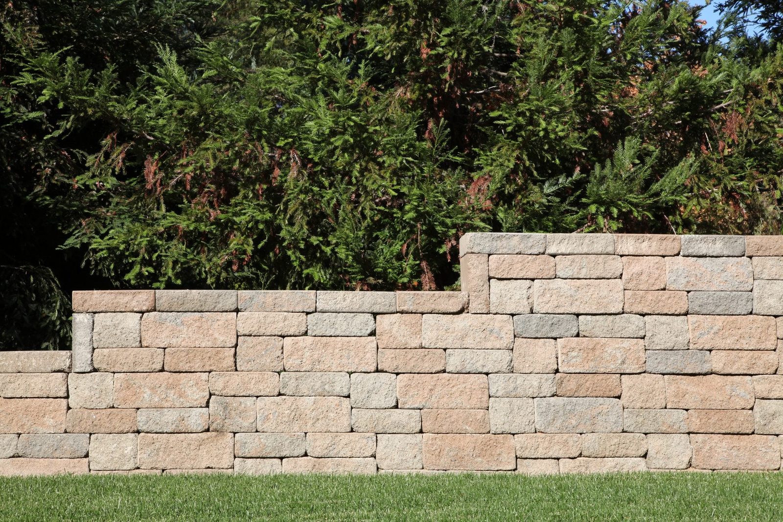 A stone wall stands firmly, showcasing toned bricks in layers, against a backdrop of lush green foliage and a well-maintained grassy area.