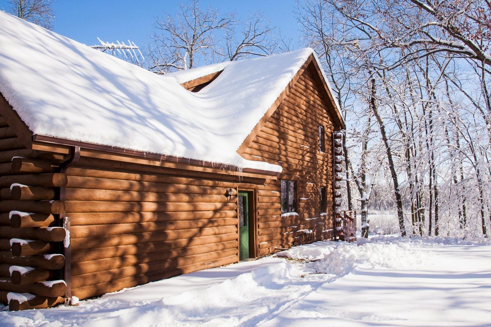 A wooden cabin is covered in snow, standing beside a snowy path, with leafless trees surrounding it under a clear blue sky.