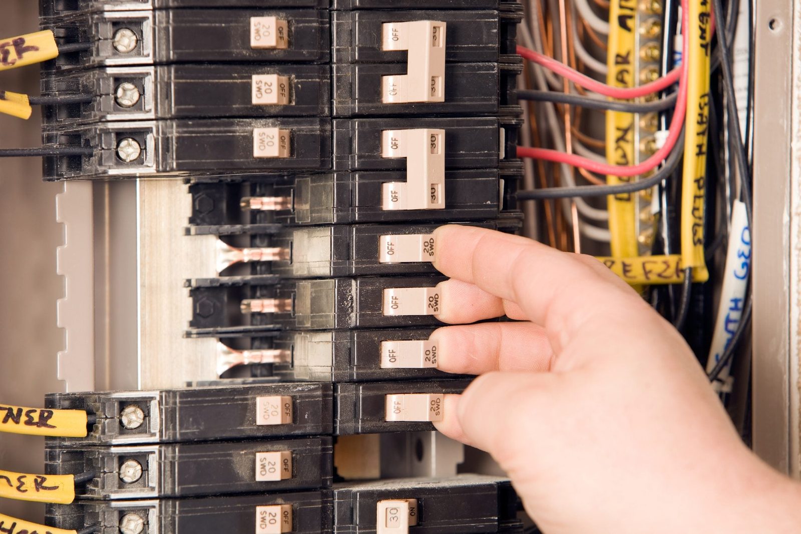 A hand is adjusting a circuit breaker inside an electrical panel, surrounded by various wires and additional breakers, indicating a maintena